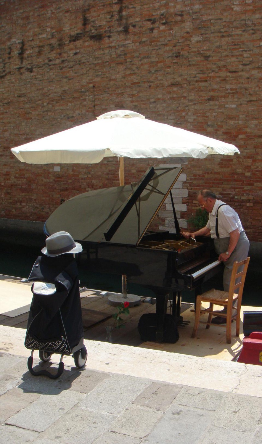 An old man tunes a grand piano placed on a small boat sitting on the waters cutting through the Venetian Ghetto. The Ghetto is a beautiful part of Venice that serves as the center of Jewish life, characterized by multiple synagogues, kosher restaurants, and Judaica shops. The community exists in a peaceful and quiet atmosphere as people can walk alongside the water, observing the ancient buildings as well as the people who currently live there.