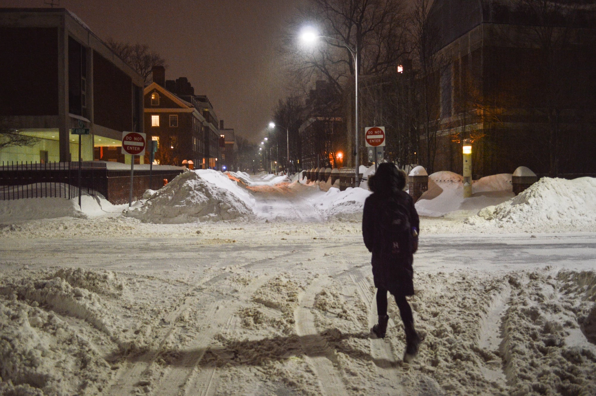 A student walks down Plympton St. on Monday evening, flanked by growing piles of snow. Amid forecasts of more wintry weather, administrators made the decision to suspend most operations at the University on Tuesday. 