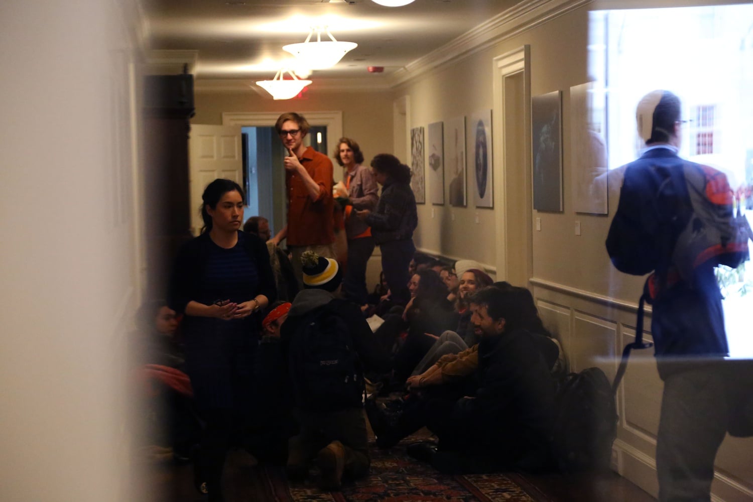 Divest Harvard member Miles Hewitt '17 gives a thumbs up as University personnel, reflected in a window, assess the situation during a protest in Massachusetts Hall on Thursday morning. 