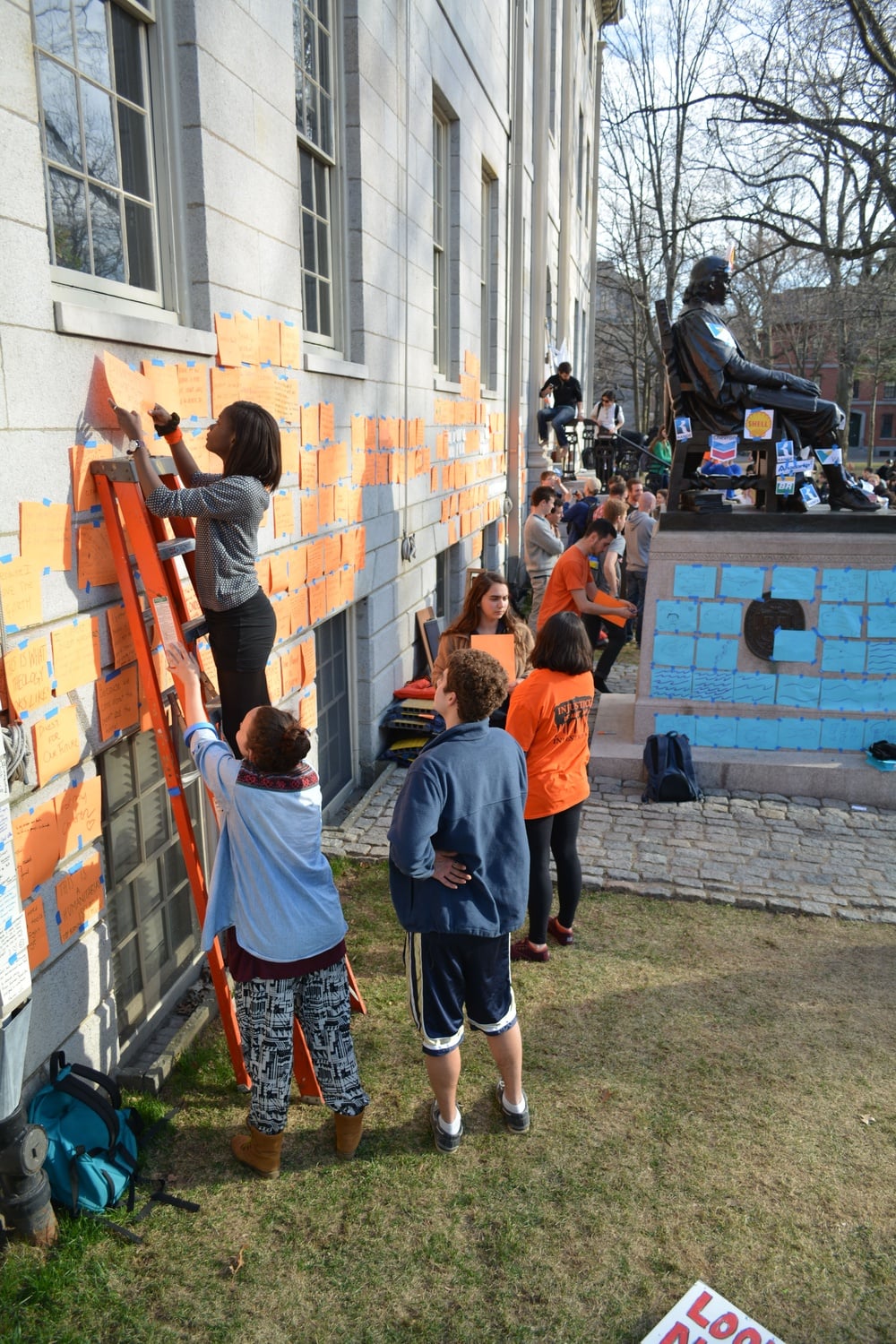 While other protesters lined up along the perimeter of University Hall, a student hangs up messages supporting the Divest movement written on orange sheets of paper. In the background, pictures of oil company logos are taped on the statue of John Harvard.
