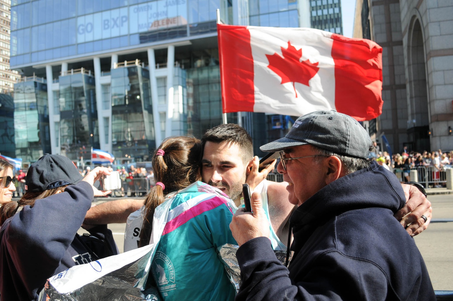 A participant embraces a friend on the sideline as he approaches the end of the Boston Marathon route. A Canadian flag - one of the many flags displayed near the finish line - billows in the wind behind. 