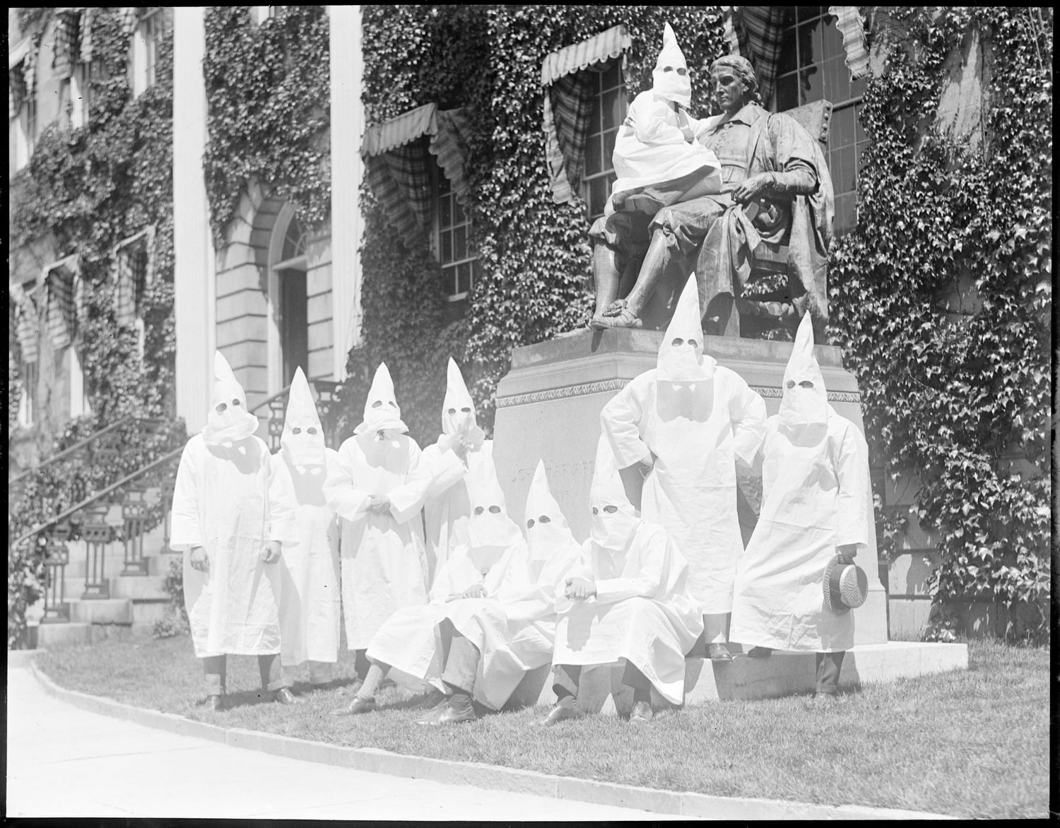 Harvard Klansmen pose for a second shot at commencement in 1924. One Klan member, sitting in the lap of the John Harvard Statue, with his body facing the statue turns his head towards the camera.