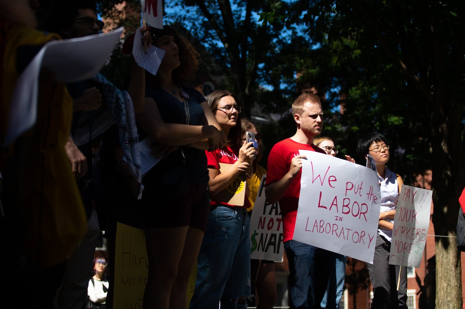 A HGSU-UAW protestor holds a sign that reads "We put the LABOR IN LABORATORY" in front of the John Harvard Statue Thursday afternoon.