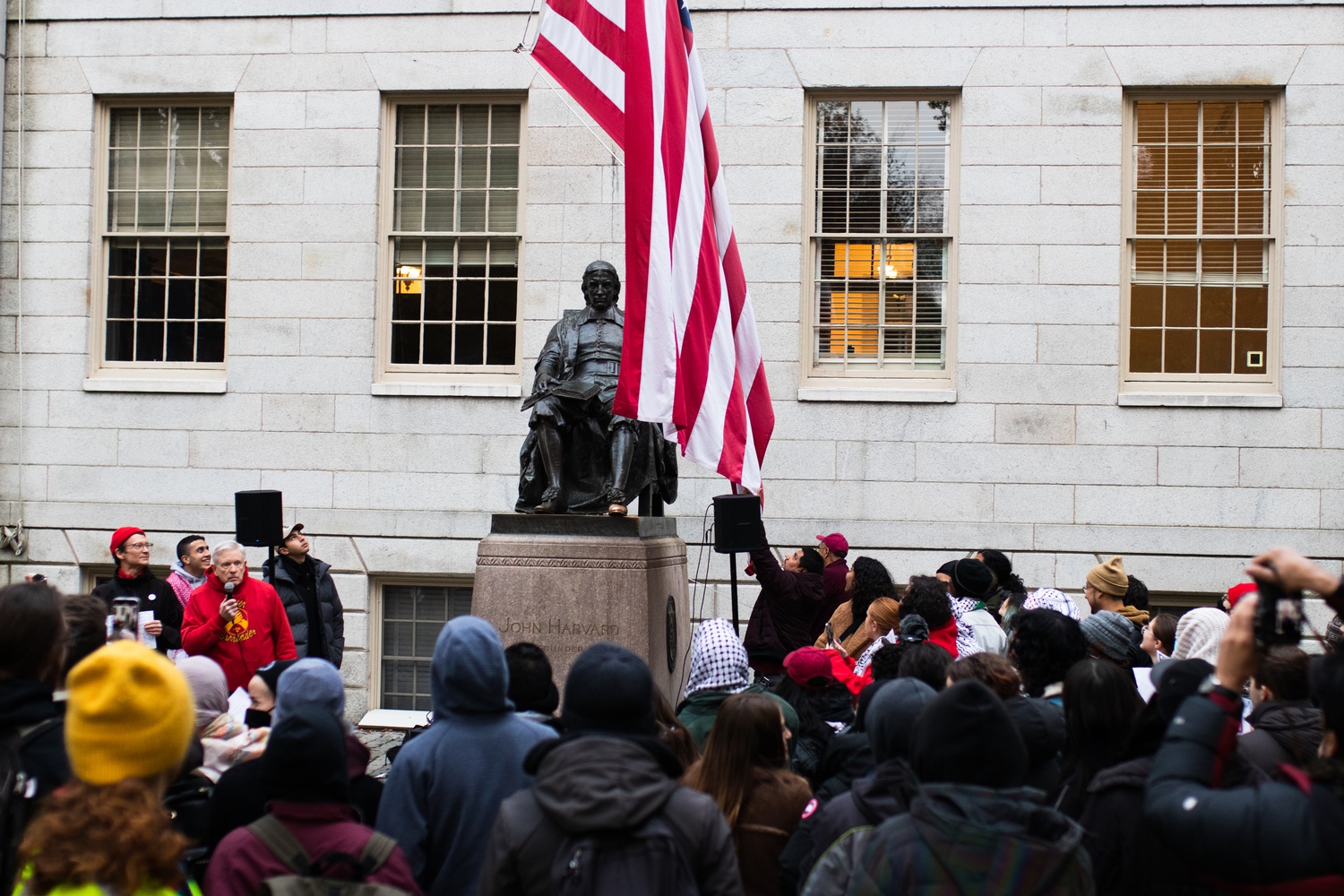 Protesters look on as Harvard personnel remove the American flag from the front of University Hall, as part of daily Harvard routine.