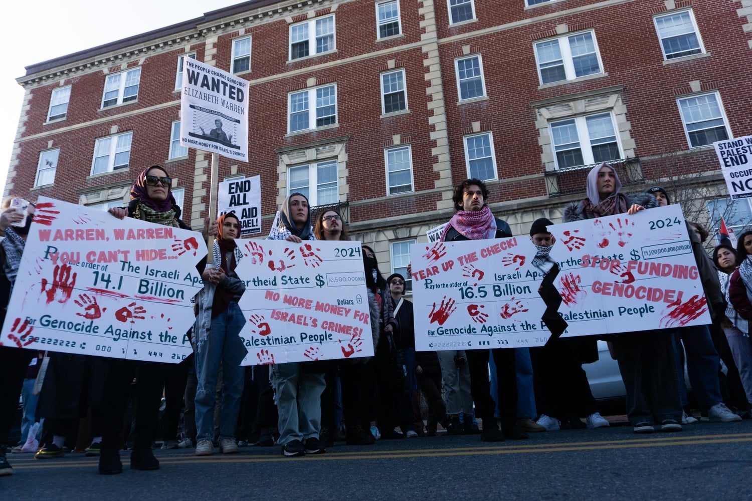 Protesters hold posters drawn as checks made out to "The Israeli Apartheid State" for "Genocide Against the Palestinian People." Chants such as "No more money for Israel's crimes!" and "Warren, Warren you can't hide..." are written in red letters over the checks.