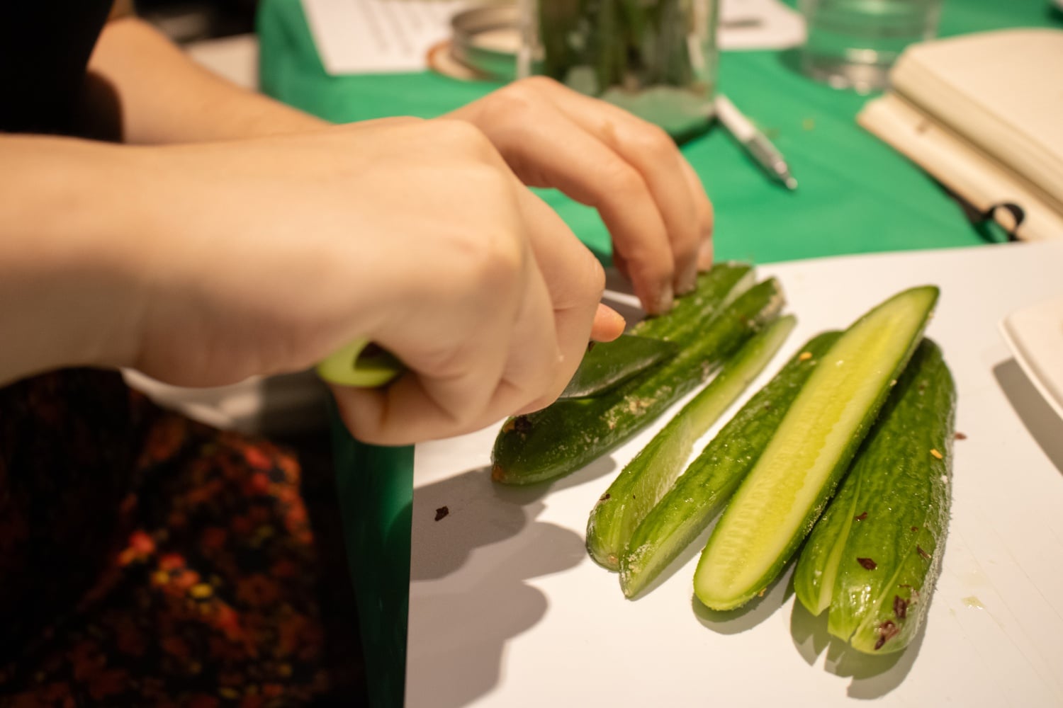 There are buckets of cucumbers in an ice bath, stacks of mason jars, and bags of green beans. Each person measures mustard seed, red pepper flakes, and heaping tablespoons of salt.