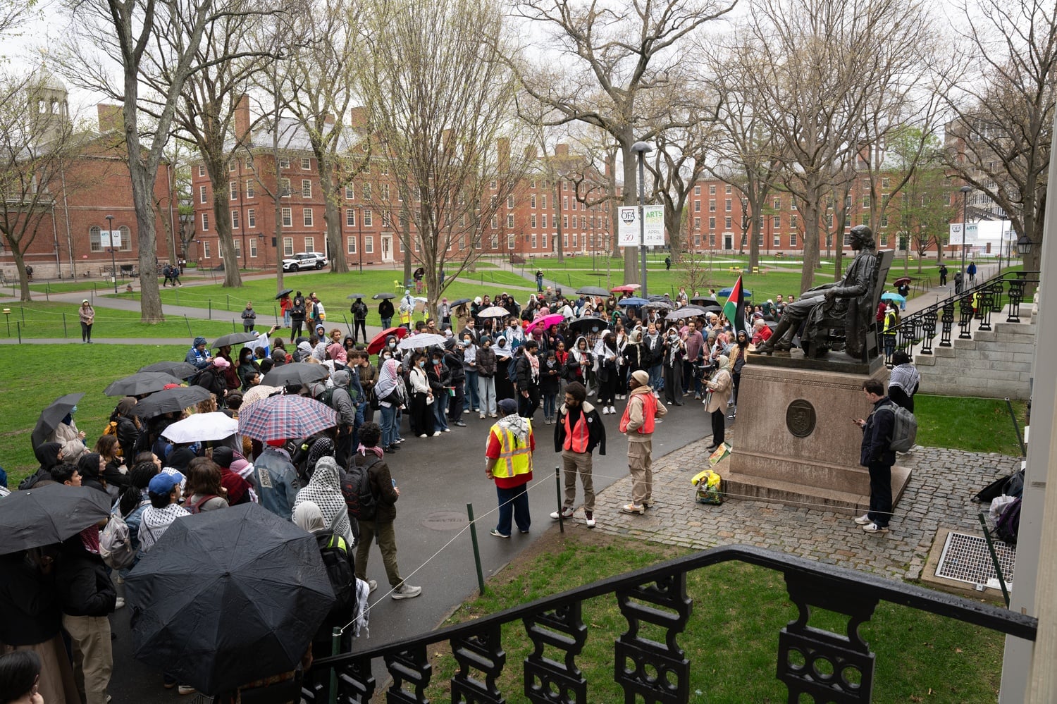 A group of protesters stand in the rain in front of the John Harvard statute in Harvard Yard.