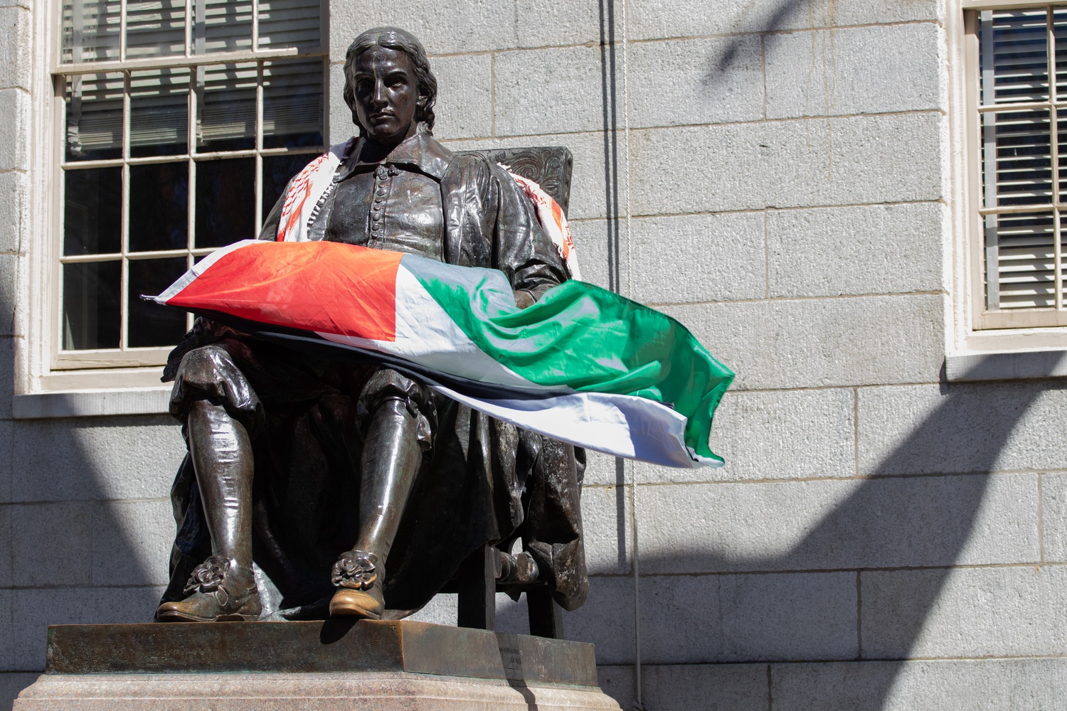 A pro-Palestine protester climbed and draped a Palestinian flag over the John Harvard statue Thursday morning. Encampment members cheered the display after sleeping overnight in Harvard Yard.