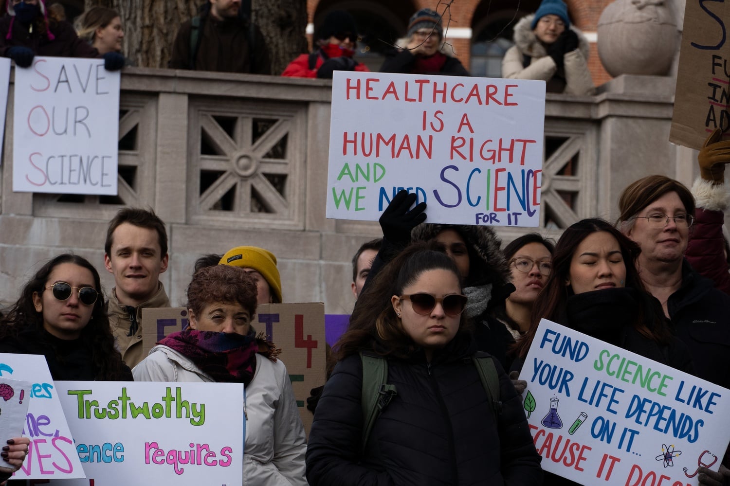 Demonstrators at the Stand Up for Science rally on Friday hold signs on the Robert Gould Shaw Memorial in front of the Massachusetts State House.