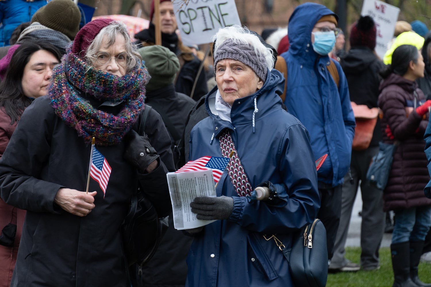 Two protesters, clad in knitwear and raincoats, hold American flags at Saturday’s rally on Cambridge Common.