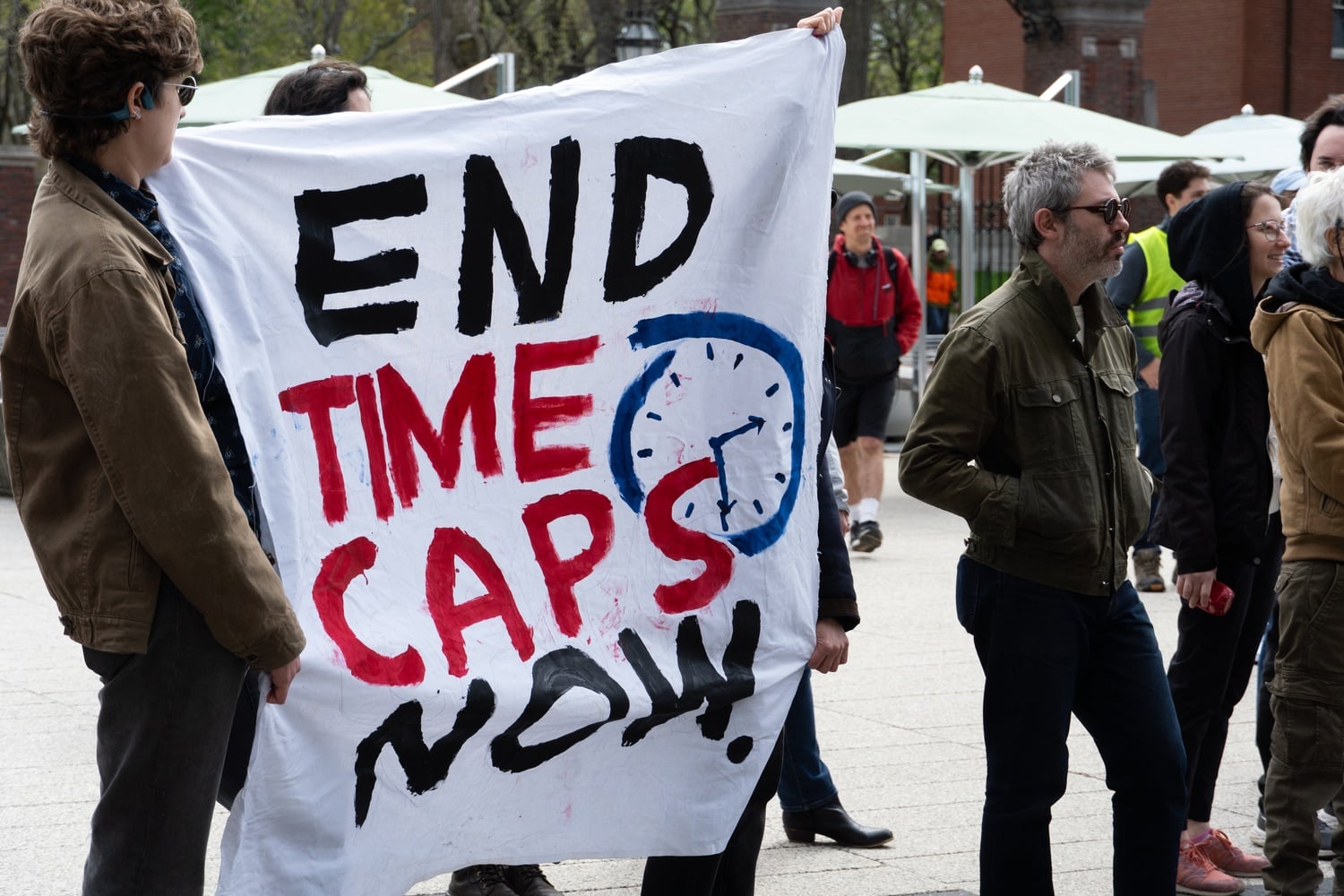 Rallygoers hold up a sign calling for an end to time caps — which limit the appointment of certain non-tenure track faculty to three, five, or eight years — at a Sunday picket in the Science Center Plaza. The rally was organized by seven campus unions and three affiliated groups to call on Harvard to protect non-citizen workers and resist federal funding cuts.