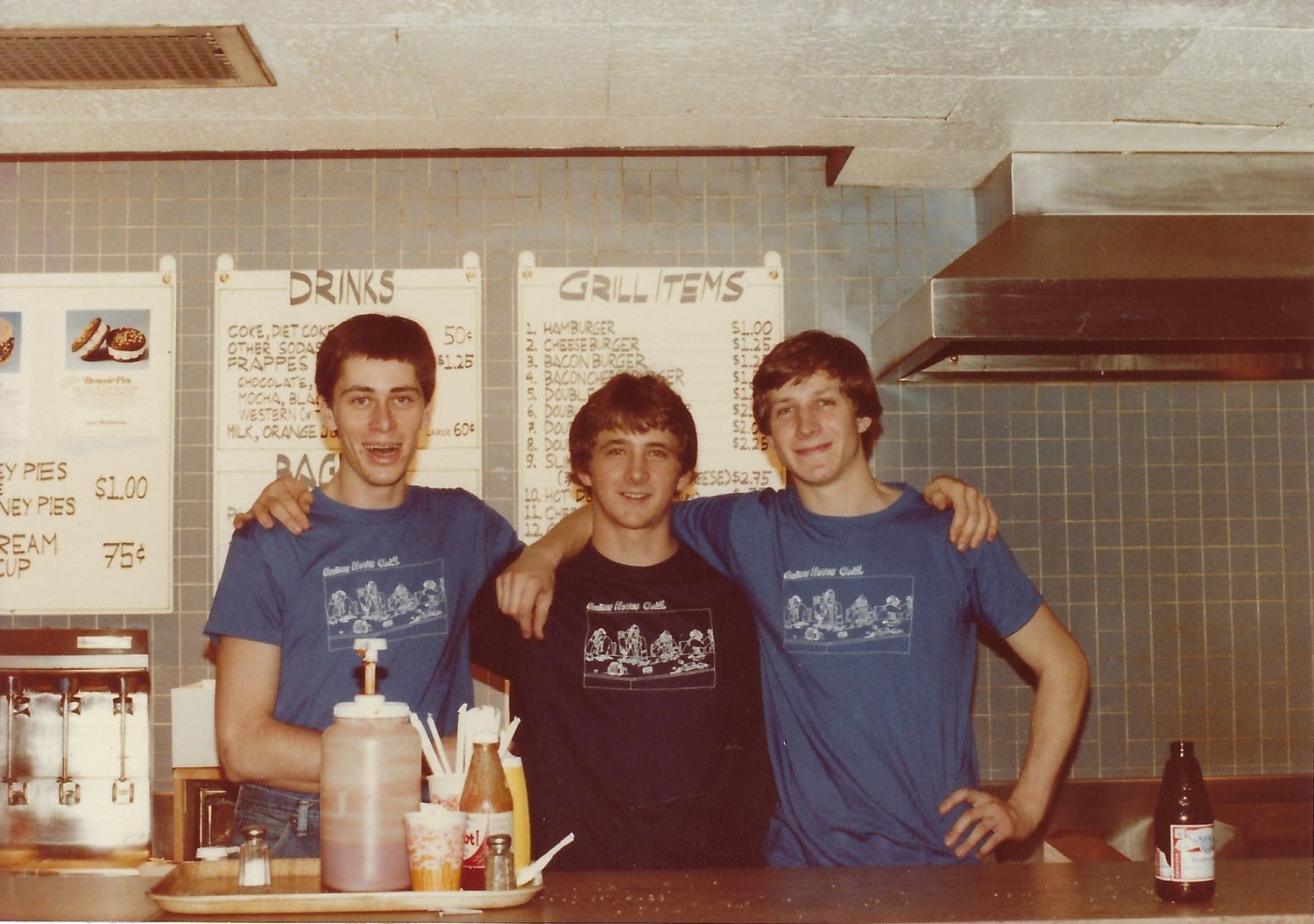 The triumvirate standing in front of the Quincy Grill (from left to right, Paul M. Manina ’84, John M. Lutz ’84, and Peter S. Kirby ’84). A beer bottle sits on the counter.