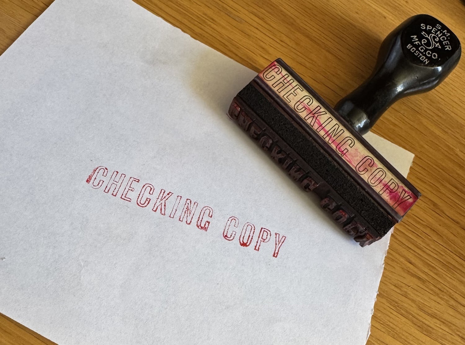 In a pile of clutter, Jona P. Liu finds a wooden stamp, smaller than a palm with a black handle and a cherry-red stained body, that prints the words "CHECKING COPY."